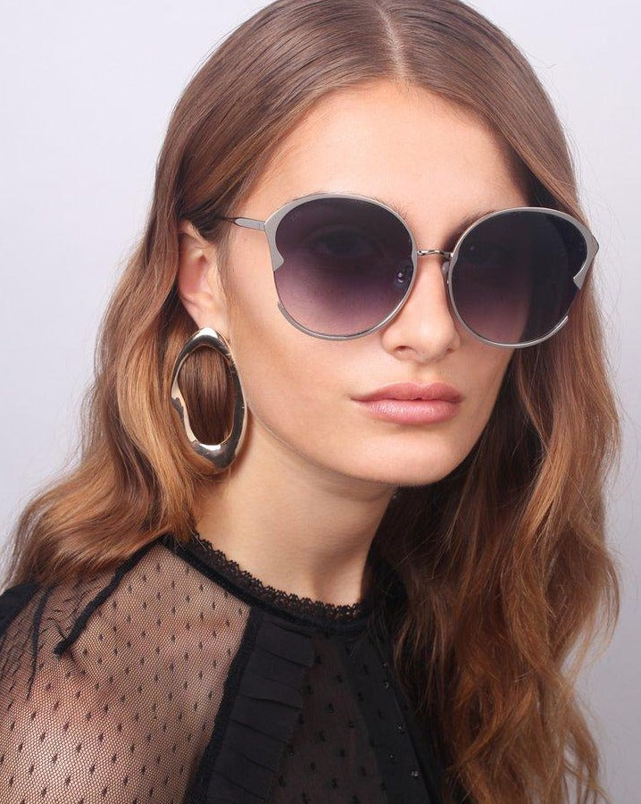 A woman with long, wavy light brown hair is wearing Alectrona large, gradient-lens handmade sunglasses by For Art's Sake® and a sheer black top adorned with delicate polka-dot detailing. She has a neutral expression and is accessorized with oversized, 18-karat gold-plated hoop earrings. The background is plain and light-colored.