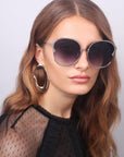 A woman with long, wavy light brown hair is wearing Alectrona large, gradient-lens handmade sunglasses by For Art's Sake® and a sheer black top adorned with delicate polka-dot detailing. She has a neutral expression and is accessorized with oversized, 18-karat gold-plated hoop earrings. The background is plain and light-colored.
