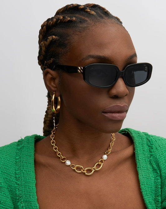A model with dark skin and braided hair wearing the For Art's Sake Bolt black sunglasses and a green cardigan with a gold and pearl necklace. 