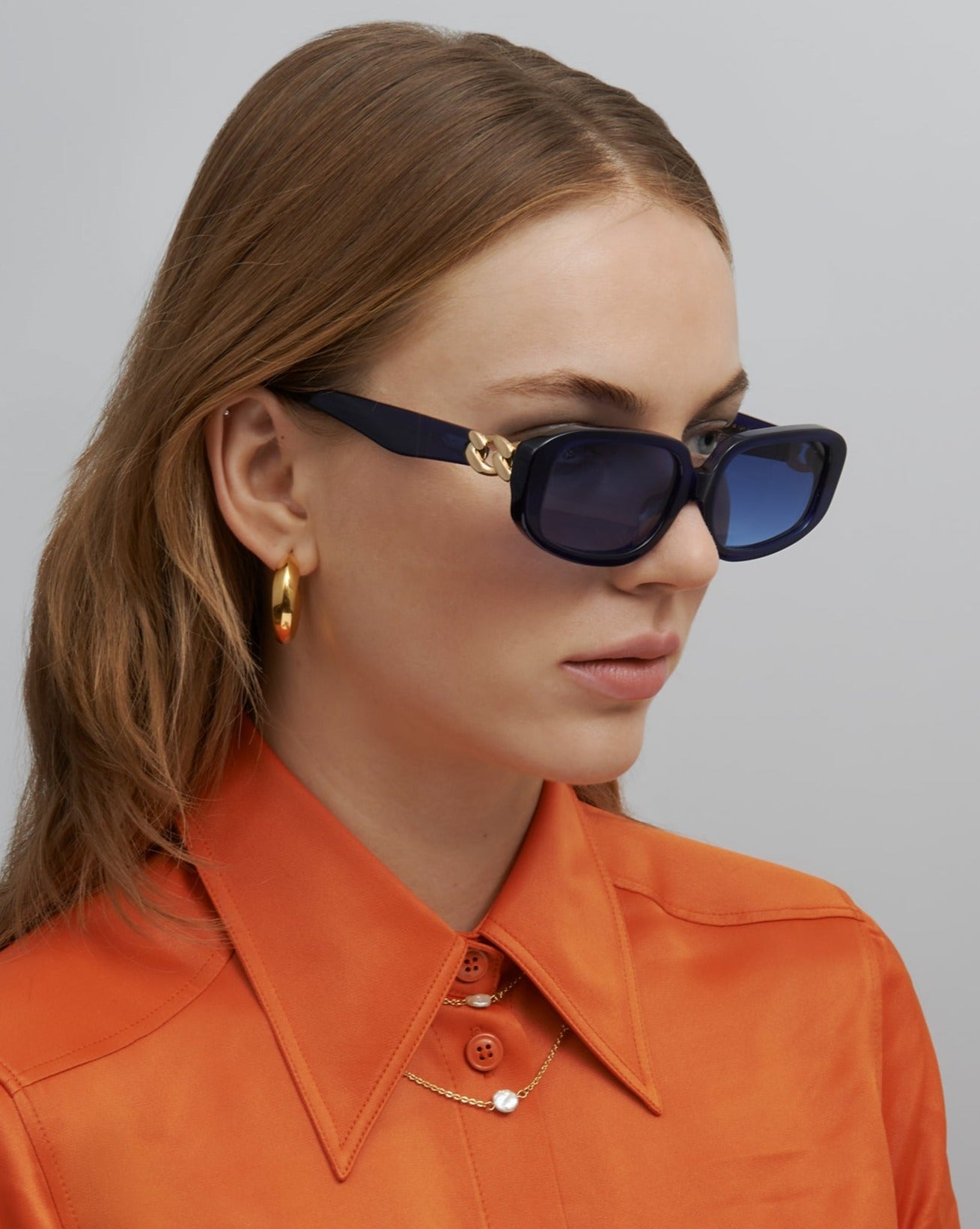 An image of a woman with Auburn hair wearing the Bolt sunglasses in Navy with an orange shirt and gold hoop earrings. 