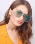A woman with long, wavy brown hair wears the Dark Eyes aviator sunglasses by For Art's Sake®, featuring stainless steel frames and nylon lenses that offer 100% UV protection. She dons a bright yellow blazer, maintains a neutral expression, and is posed against a plain background.