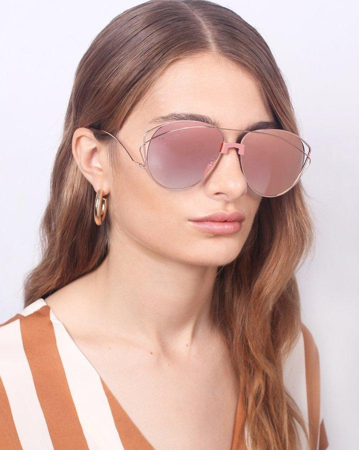 A woman with long, wavy brown hair is wearing the "Dark Eyes" sunglasses by For Art's Sake®. These large, reflective pink sunglasses feature stainless steel frames and Nylon lenses that provide 100% UV protection. She has a neutral expression and is dressed in a brown and white striped top, set against a plain white background.
