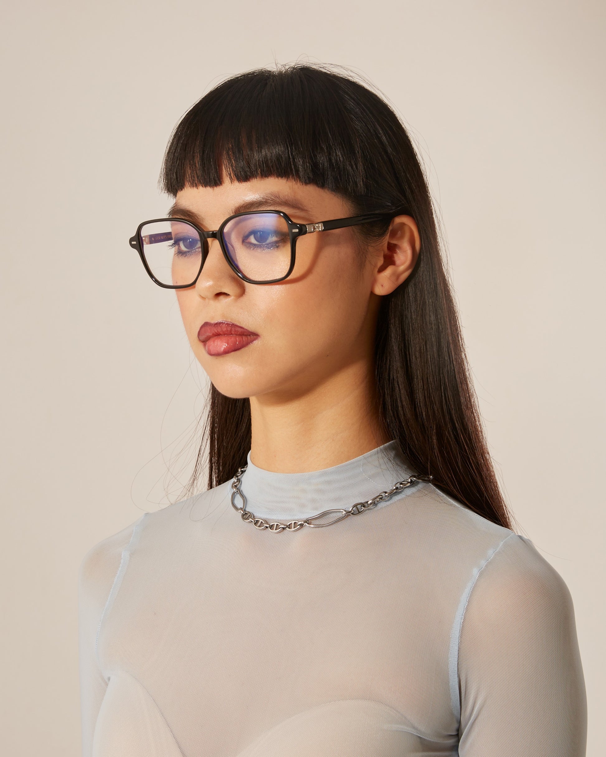 A woman with straight black hair and black cat-eye framed optical glasses, wearing a sheer, ribbed top and a chain necklace.