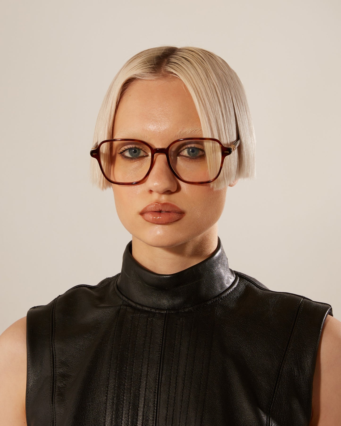 A woman with a short, blunt bob haircut wearing the Azure brown optical glasses by For Art’s Sake and a black sleeveless turtleneck, giving a serious look toward the camera against a neutral background.