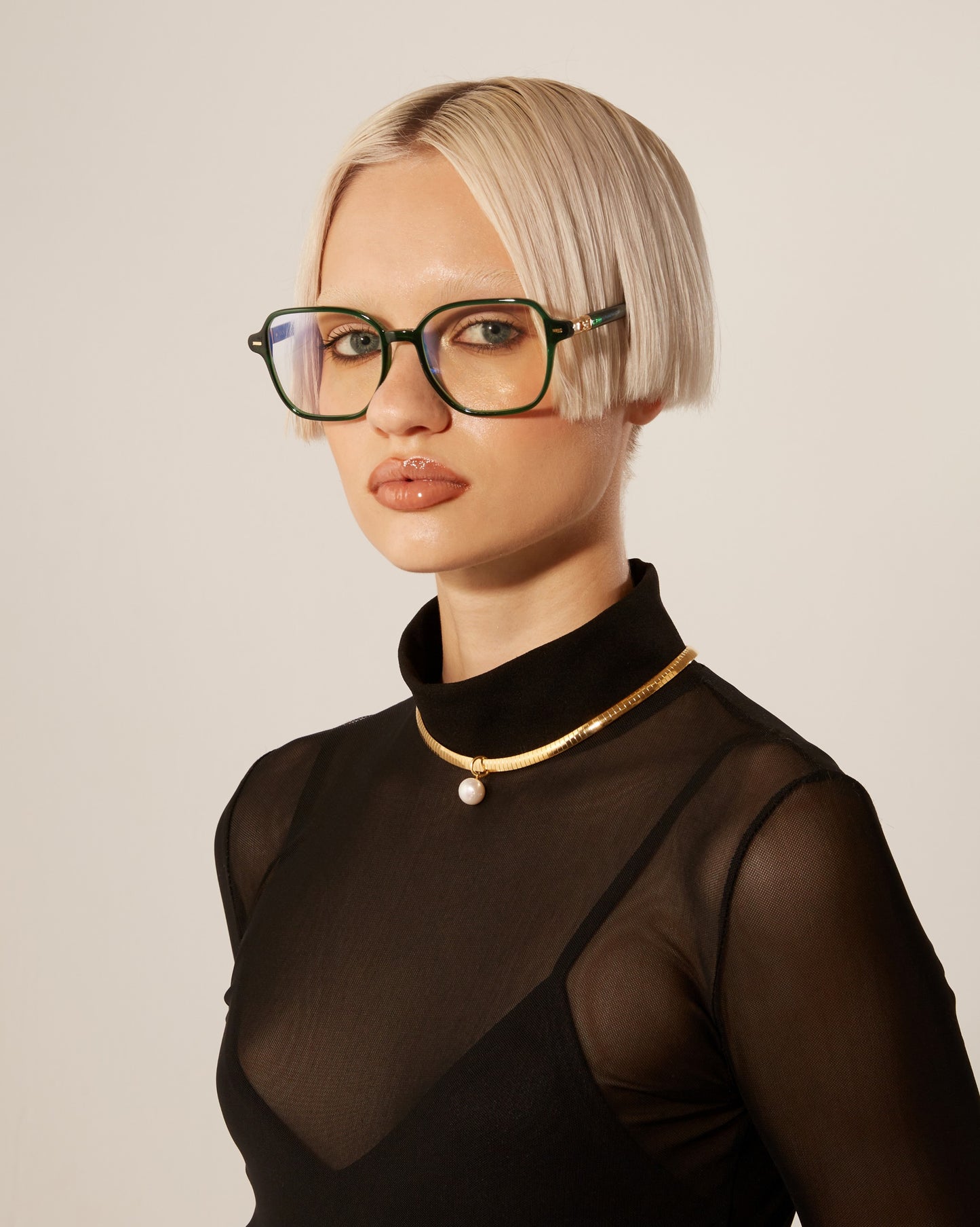 A woman with short blonde hair and green-frame optical glasses, wearing a sheer, black top and a chain gold necklace with a pearl. 