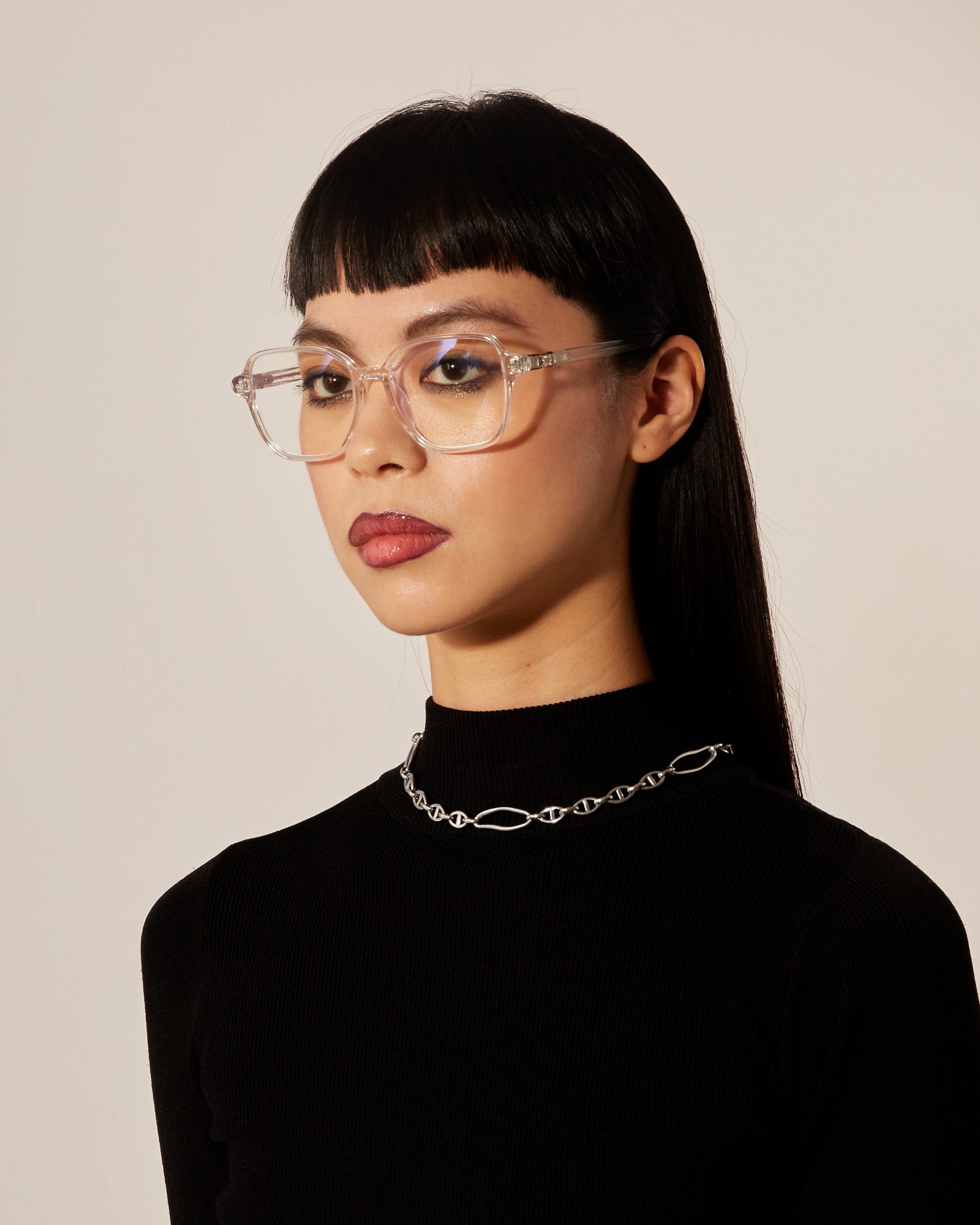 A woman with straight black hair and clear-framed optical glasses wearing a black turtleneck and a silver chain necklace. 