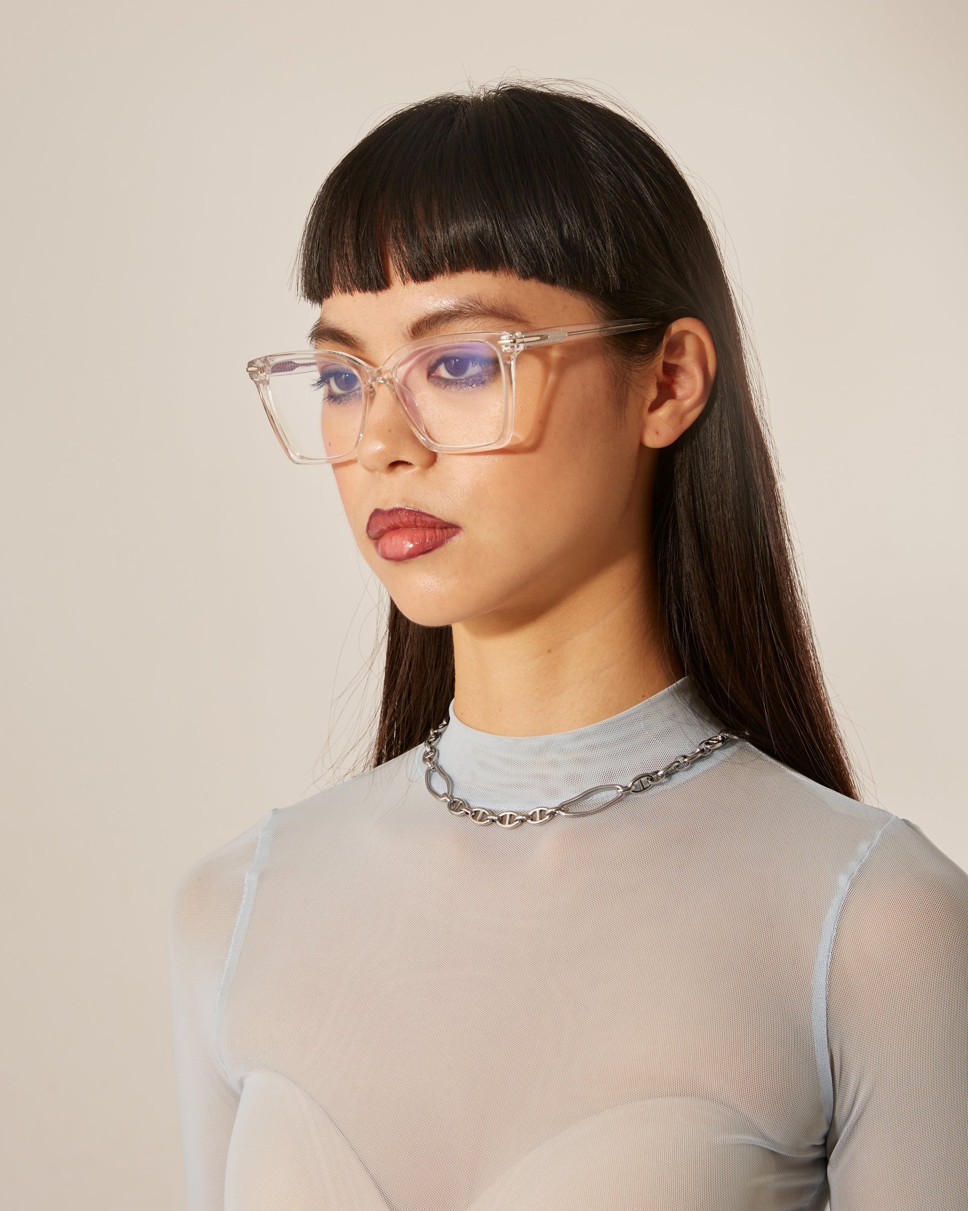 A woman with straight black hair and clear-frame optical glasses, wearing a sheer, ribbed top and a chain necklace.